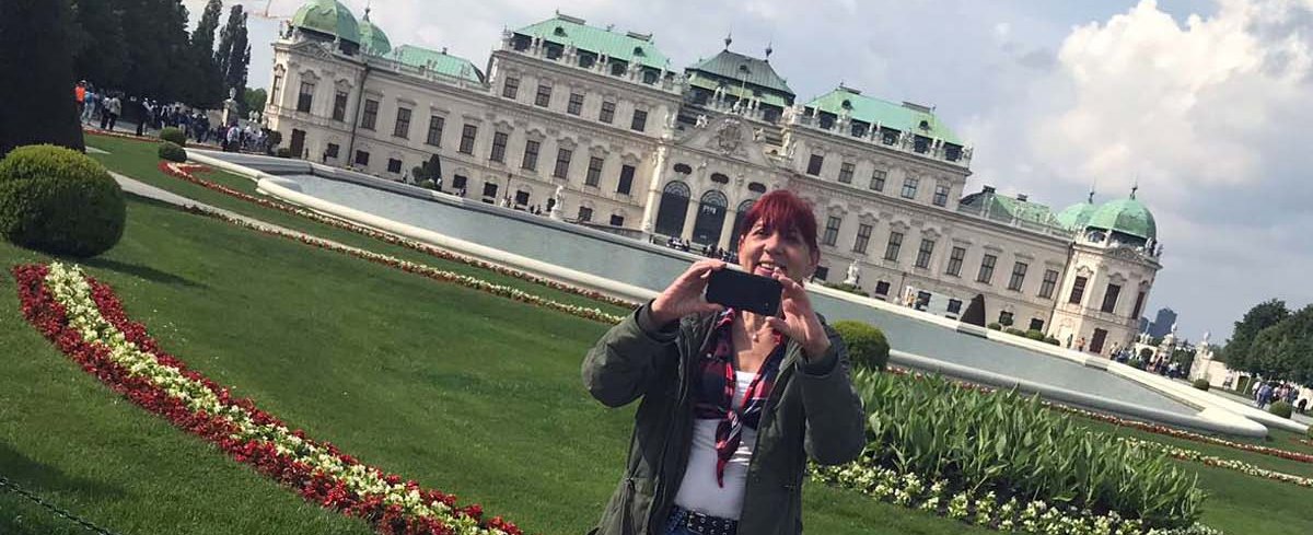 Birgit Proietto steht in einem Park, hinter ihr ist ein Schloss zu sehen.  Sie macht ein Selfie-Foto mit dem Schloss im Hintergrund.. 