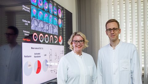 Dr. Johanna Diekmann und Dr. Tobias König stehen vor einem Fernseher an der Wand auf dem Ihre Arbeit zu sehen ist.