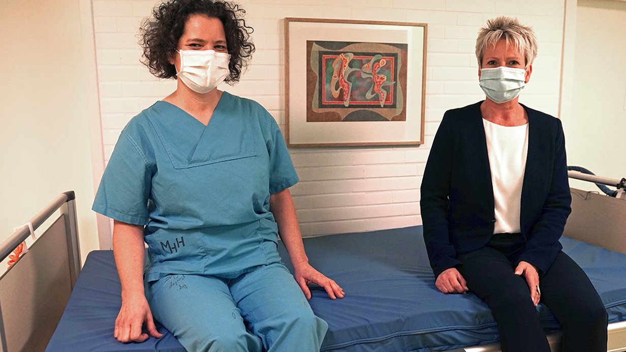 Beeke Luise Schmeling und Dr. Regina Schmeer sitzen mit Mund-Nasen-Bedeckung auf der neuen Matratze.