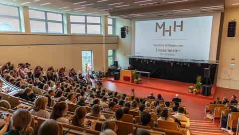 ein Foto der Erstsemester, die in einem Hörsaal sitzen. Fotografiert wurde vom oberen Bereich des Hörsaals nach unten. Ein Beamer projeziert die Worte "Herzlich Willkommen Erstsemester" an die WandWorte