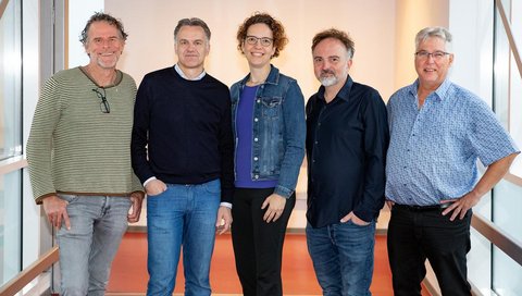 In einem Gang stehen vier Männer und eine Frau und lächeln in die Kamera.