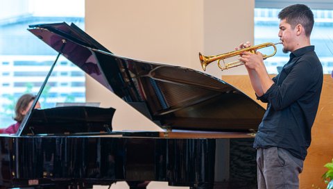 A man is playing the trumpet, behind him is a grand piano. 