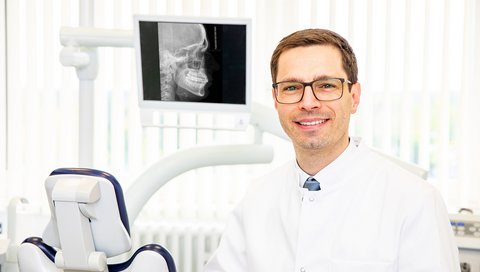 Prof. Kunz stands in front of a dental chair in the Clinical Department of Orthodontics; behind him, a monitor displays an X-ray image. 