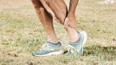 A person is holding their sore calf and wearing turquoise sneakers on green grass.