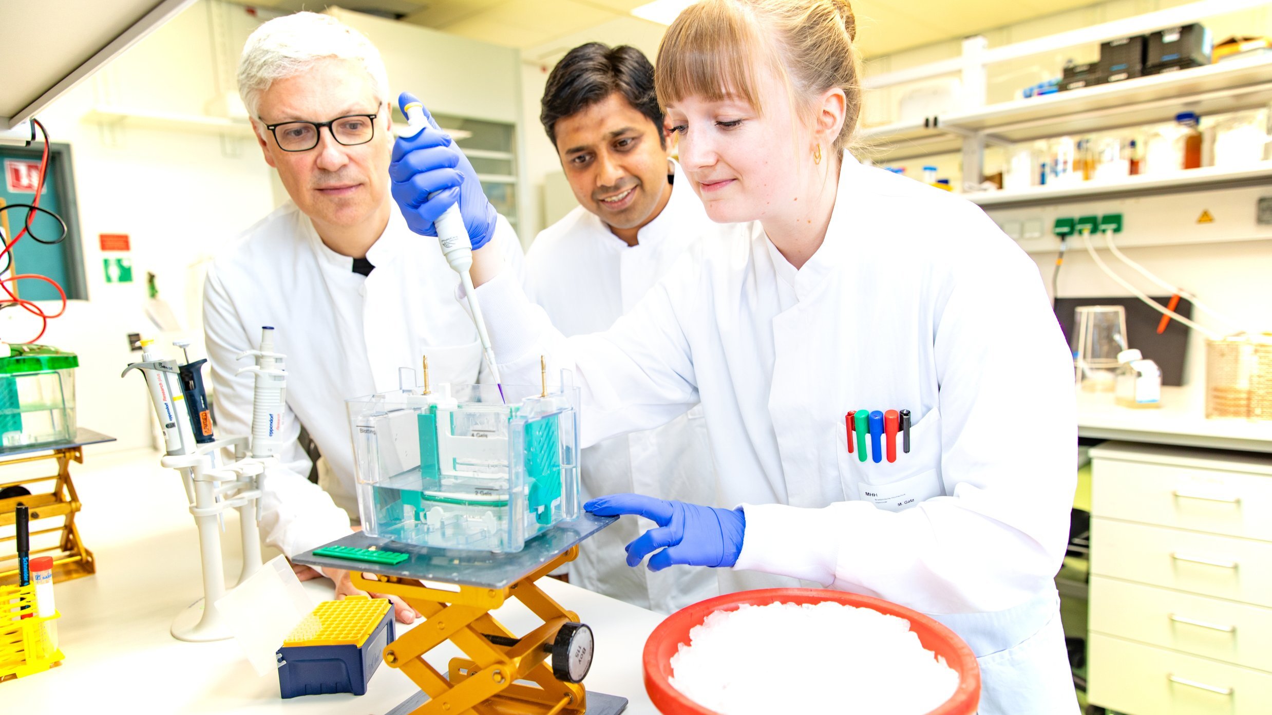 Zwei Männer im weißen Laborkittel stehen in einem Labor und schauen einer jungen Wissenschaftlerin beim Pipettieren zu. 