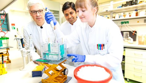 Zwei Männer im weißen Kittel stehen in einem Labor und sehen einer jungen Wissenschaftlerin beim Pipettieren zu.