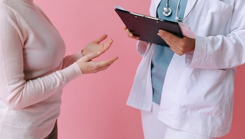 Zwei Personen stehen sich gegenüber, die Person rechts trägt einen Arztkittel und in den Händen ein Klemmbrett, die Person links hält die Hände wie bei einem Gespräch hoch vor ihrem Oberkörper.