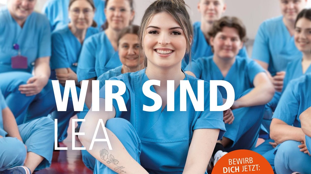 Plakatmotiv mit mehreren Pflegekräften in blauen Kasacks, die auf einem Flurfußboden knien. Im Vordergrund sitzt eine weibliche junge Pflegekraft und lächelt in die Kamera.