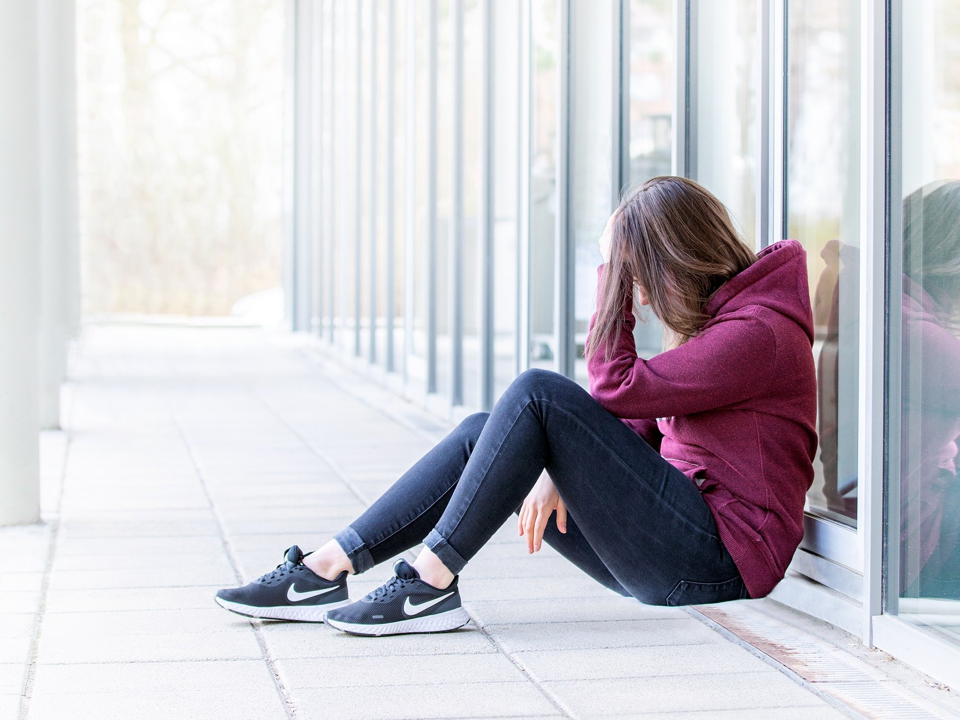 eine junge Frau mit braunen Haaren sitzt im Bild rechts and eine Glasfassade gelehnt, hat die Knie angewinkelt und stützt den Kopf mit der Hand ab