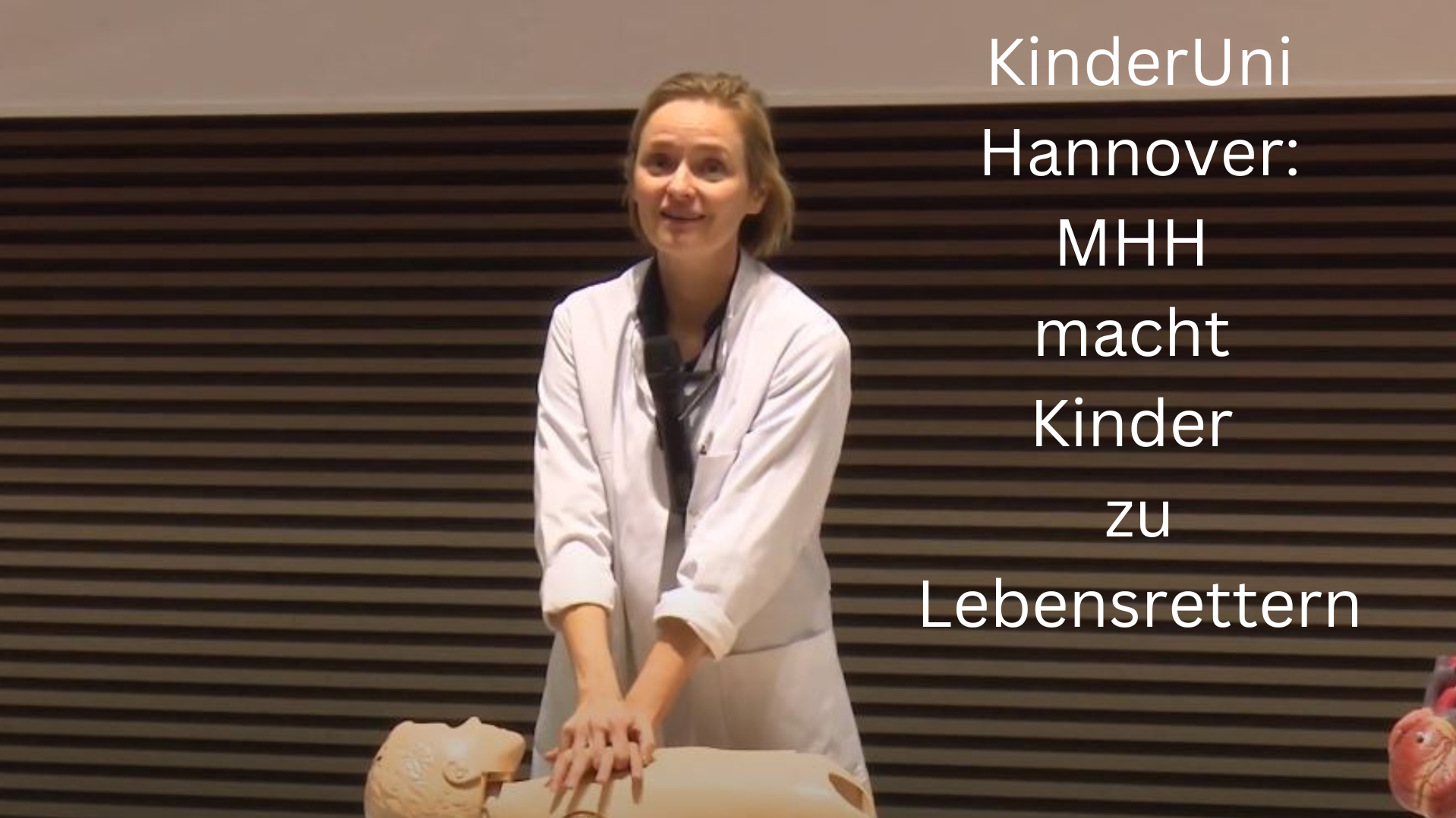 Frau Dr. Happle steht vorne auf dem Podium eines MHH-Hörsaals und demonstriert die Herzdruckmassage an einer Übungspuppe. 