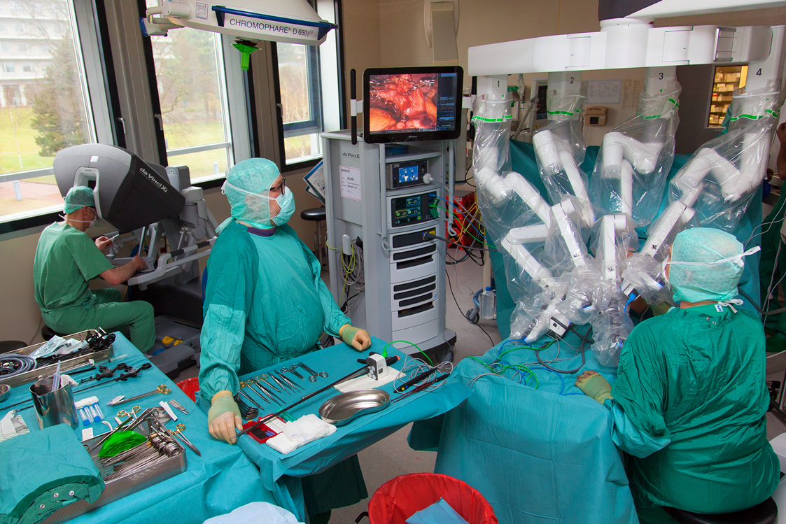 Blick in den OP-Saal auf den OP-Monitor und die Arme des OP-Roboters OP-Saal mit zwei OP-Kräften stehend am abgedeckten Patienten, im Hintergrund Chirurg an der Konsole, Monitor zeigt Körperinneres