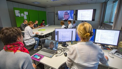 Das Expertenteam, bestehend aus verschiedenen Personen, sitzt in einem Raum, auf Bildschirmen sind Unterlagen zu sehen, eine Person ist über Videokonferenz dazu geschaltet.