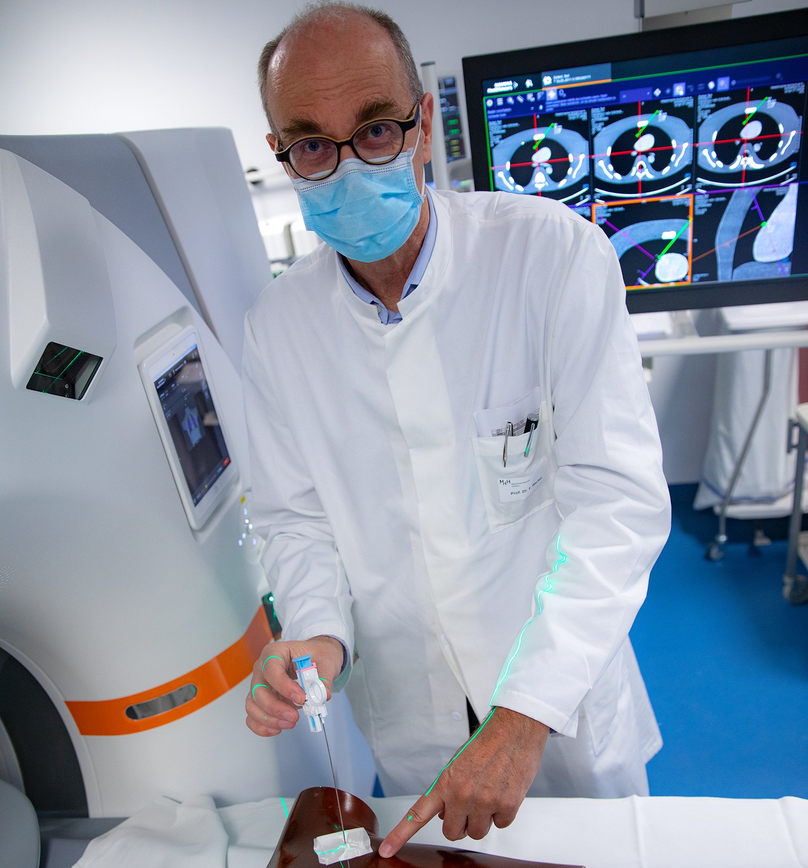 The picture shows Professor Wacker at the new CT machine with laser navigation. He uses an artificial model to demonstrate how green laser beams mark exactly the right puncture site for the needle.