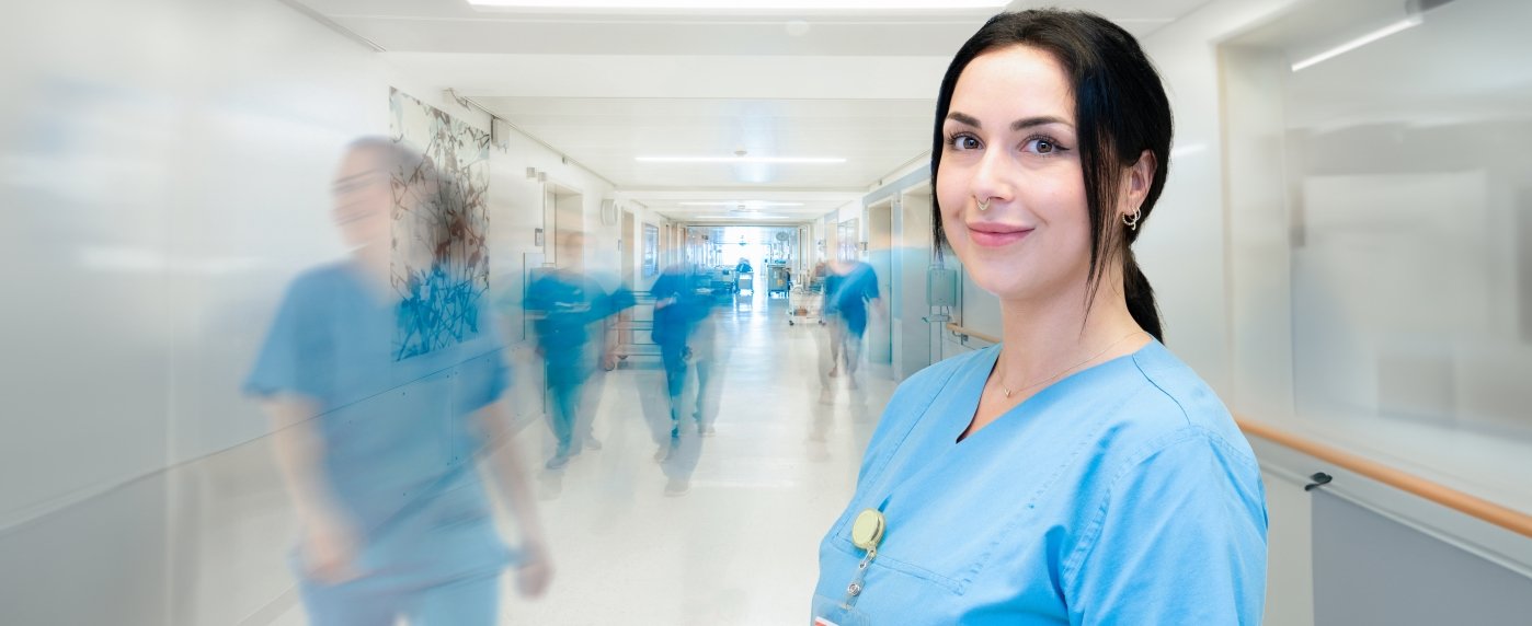 Eine Pflegekraft in blauem Kasack steht in einem Stationsflur, blickt leicht lächelnd vorn in Kamerarichtung. Im Hintergrund sind andere Pflegekräfte in Zeitraffer verschwommen zu sehen. 