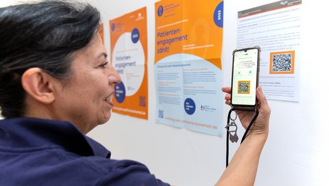 Dr. Maria Cartes stands in front of a poster, which shows the QR Code. She scans it with her smartphone.