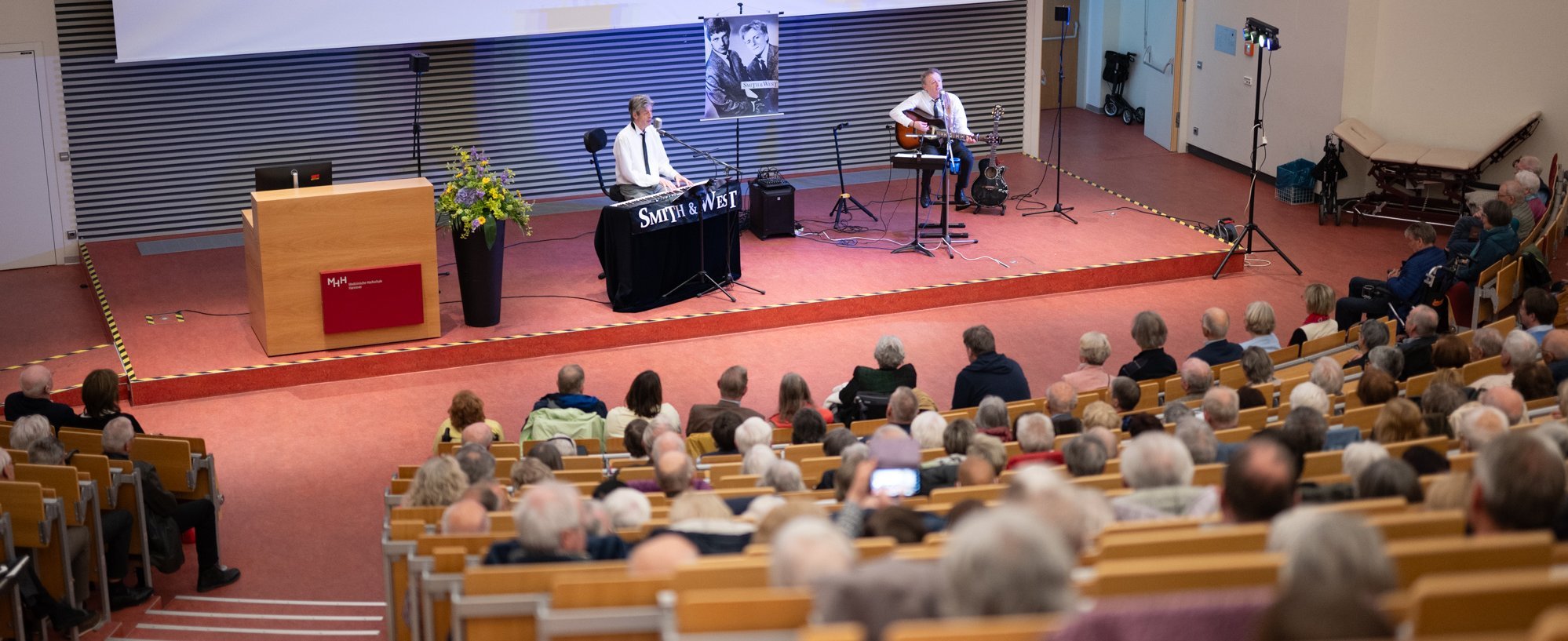 Im Hörsaal der MHH sitzen viele Menschen, vorne auf der Bühne spielt ein Mann Gitarre, ein anderer Keyboard und beide singen in eine Mikrofon. 