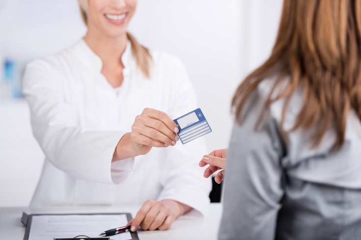 Frau in weißem Kittel überreicht Person eine Krankenversichertenkarte