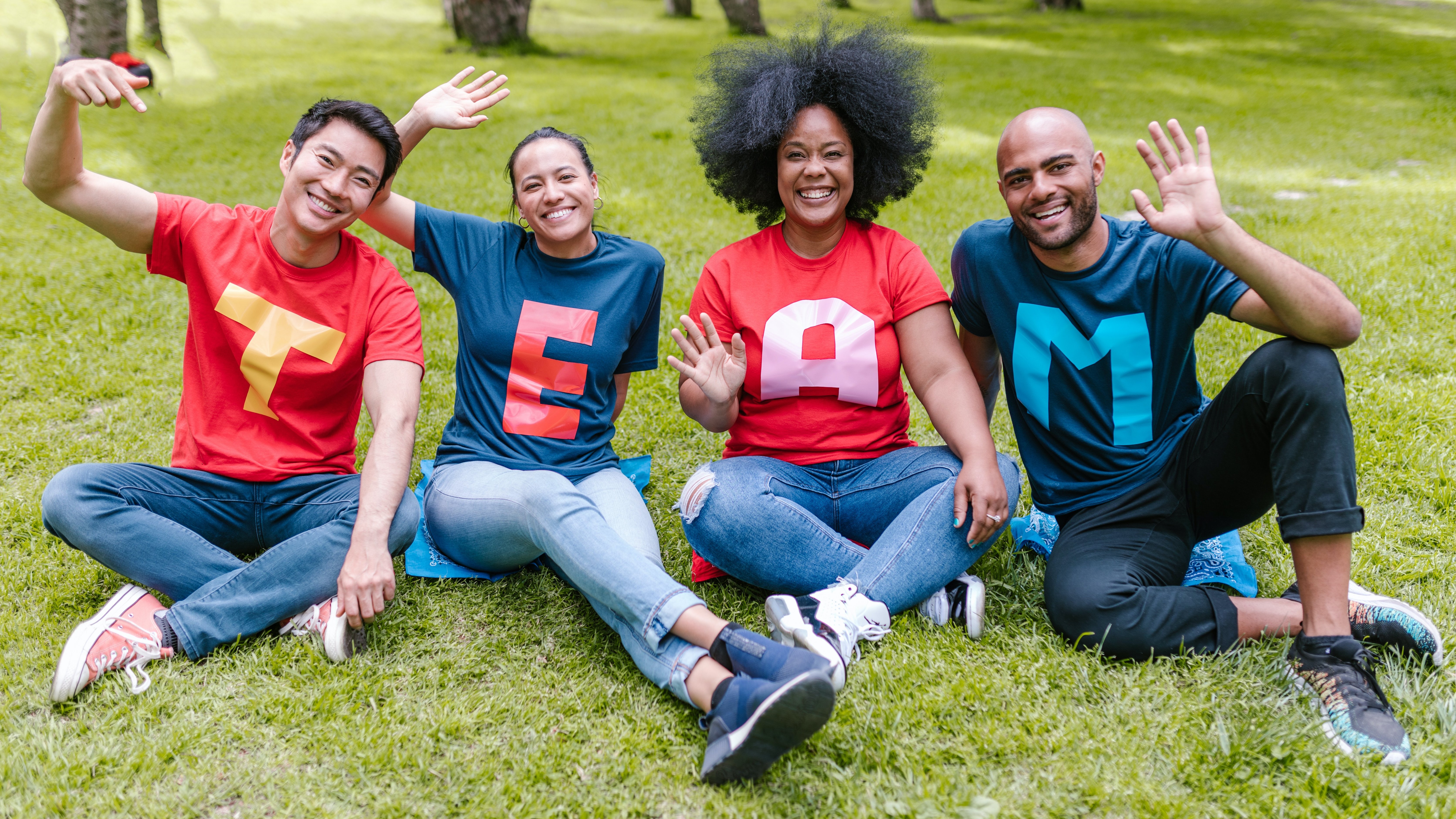 The picture shows for people sitting in a park laughing and waving at the camera. On each of their shirts is a letter, together they spell out the word "Team".