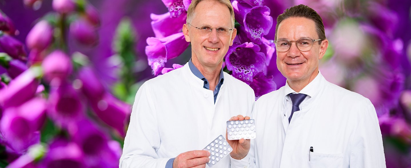 Zwei Männer im weißen Kittel stehen vor einem Foto mit rotem Fingerhut und halten Tabletten-Blister in der Hand.