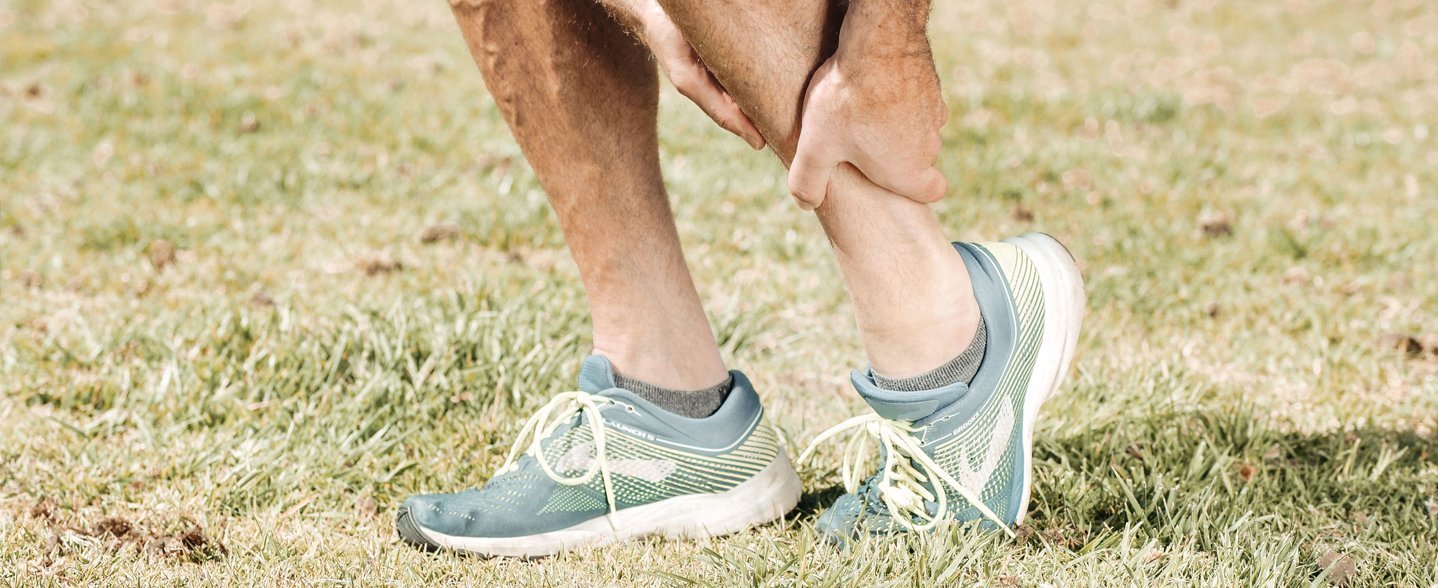 A person is holding their sore calf, wearing turquoise sneakers on green grass.
