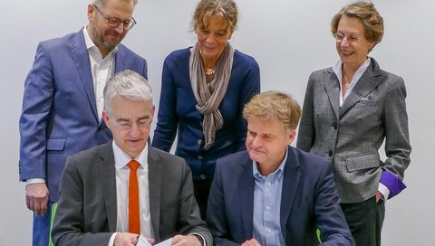 Fünf Personen stehen und sitzen an einem Tisch, auf dem ein Dokument liegt.