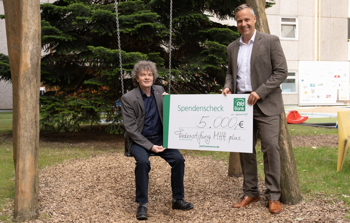 Dr. Schenke sitzt auf einer Schaukel im Innenhof der Kinderklink, Neben ihm steht Torsten Krieger von der PSD Bank. Gemeinsam halten sie den Spendenscheck hoch.