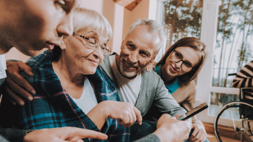 Two man and two woman, one is an elderly woman, sit together and look at a phone together.
