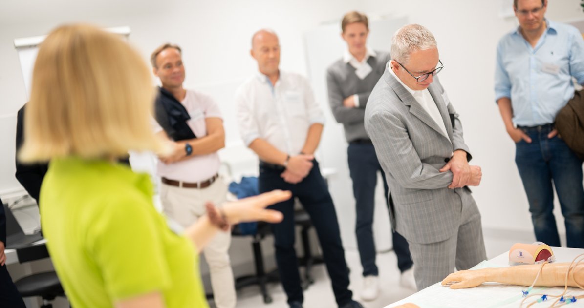 Eine studentische Tutorin im Vordergrund erklärt der Gruppe eine Reihe von Modellen, die auf einem Tisch liegen.