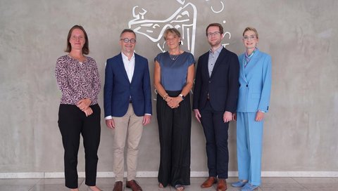 Fünf Personen stehen vor einer Wand mit einem kunstvollen Schriftzug und einem grafischen Motiv. Die Gruppe blickt lächelnd in die Kamera.