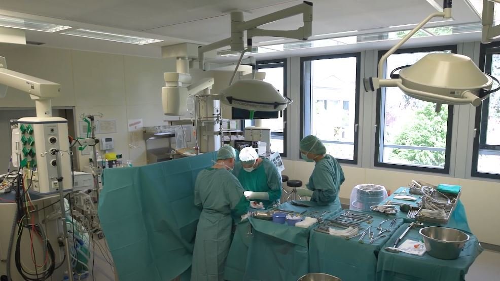 Blick von oben in einen OP-Saal mit drei Personen in OP-Schutzkleidung, die an einem OP-Tisch stehen. 