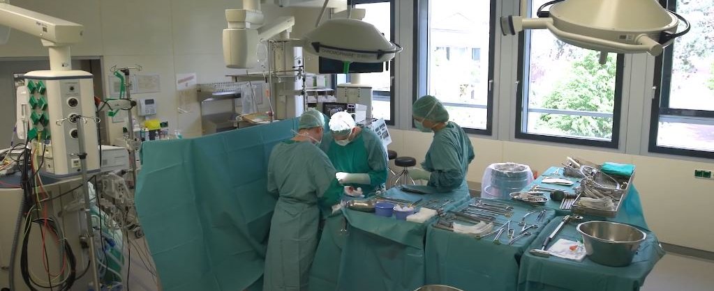 Blick in einen OP-Saal, in dem drei Menschen in grüner OP-Schutzkleidung an einem OP-Tisch stehen. 