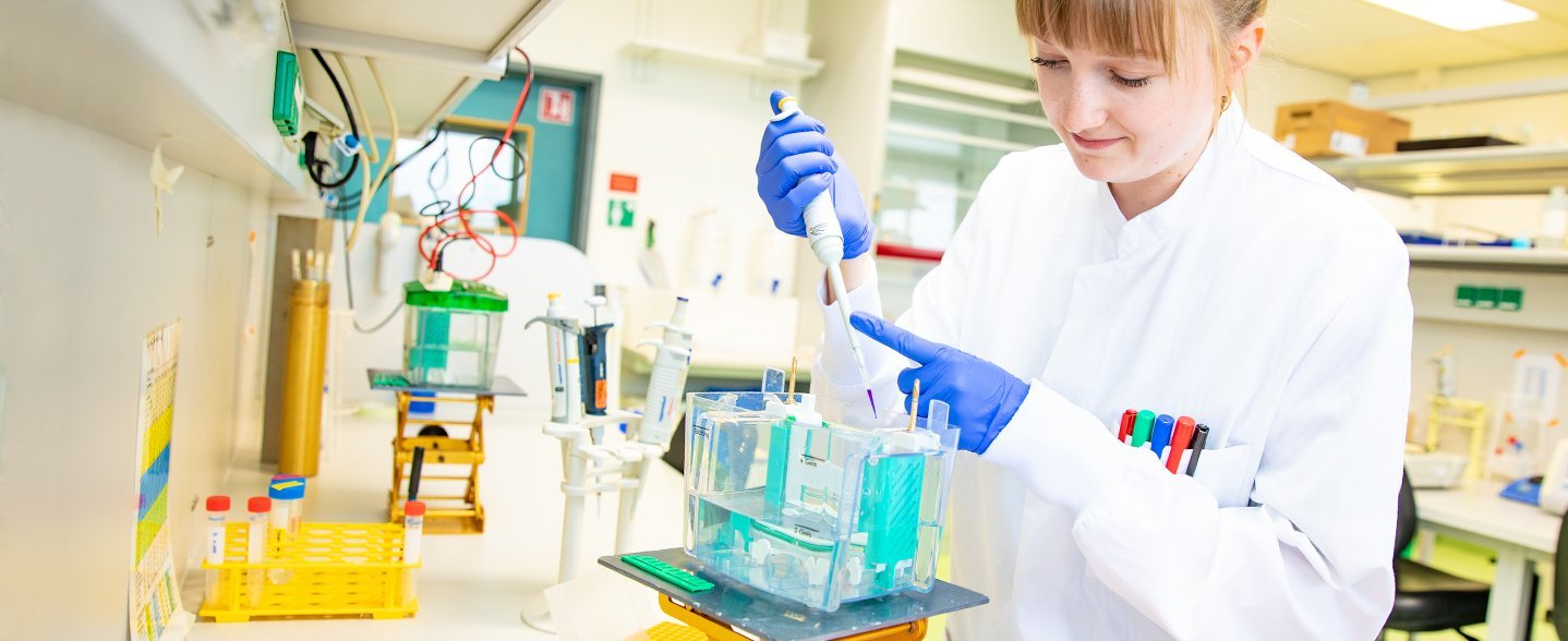 eine junge Wissenschaftlerin im weißen Kittel steht im labor und pipettiert