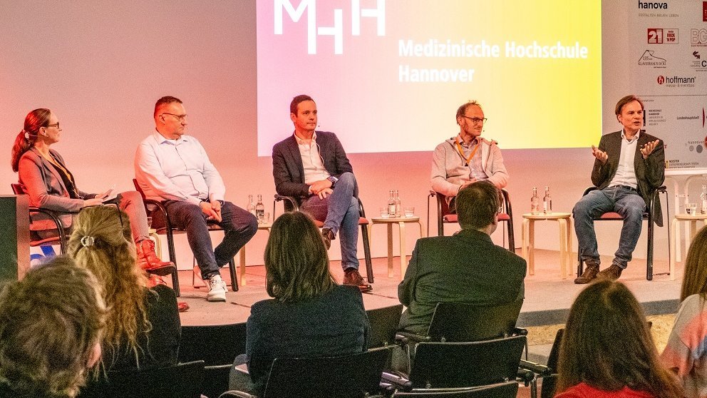 Auf einem Podium im aufhof sitzen fünf Personen. Vor ihnen sitzen Menschen in mehreren Stuhlreihen. 