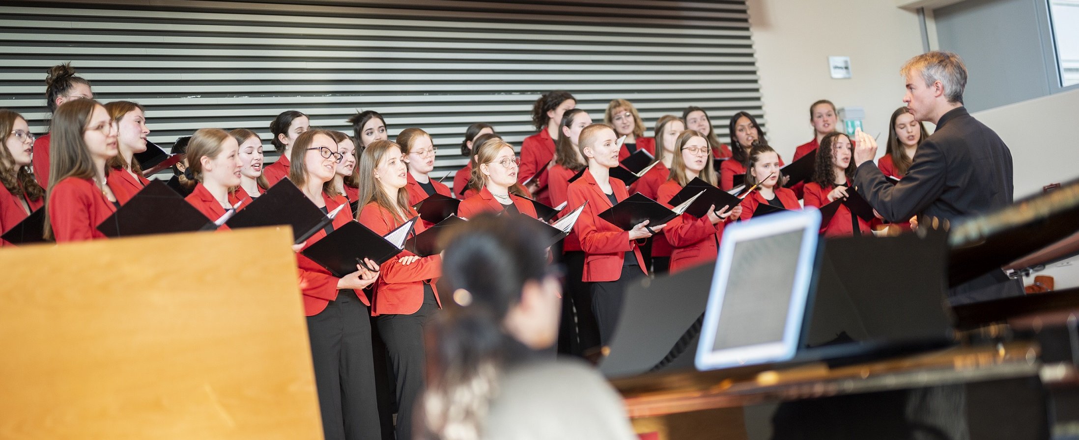 Der Mädchenchor singt im Hörsaal F der MHH. Die jungen Frauen tragen rote Blazer und schwarze Hosen. Vor ihnen steht Leiter des Chors, Professor Felber.