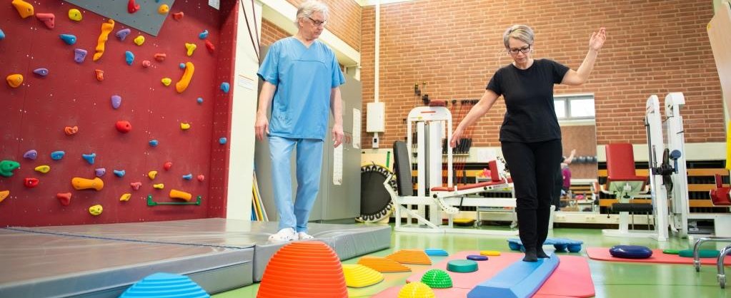 Der Rehabilitationsraum der MHH.  Eine Patientin balanciert auf einem trapezförmigen Gegenstand. Neben ihr läuft eine anleitende Person.