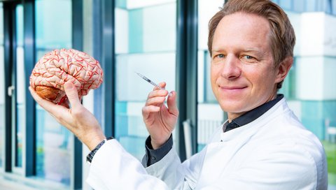 Professor Dr. Tillmann Krüger hält das Modell eines menschlichen Gehirns und eine Spritze in den Händen.