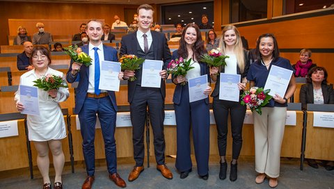 Die Promotionspreisträgerinnen und Promotionspreisträger (von links): Dr. rer. nat. Fangfang Chen, Dr. rer. nat. Alexander Bollenbach, Dr. med. Tammo Lambert Tergast, Dr. med. Nora Möhn, Dr. rer. nat. Anna Christina Dragon und Dr. rer. biol. hum. Rizky Indrameikha Sugianto stehen in einem Hörsaal, hinter ihnen die Sitzbänke.