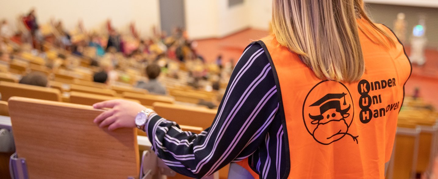 Eine Person mit orangener Weste blickt von oben in einen Hörsaal. Auf der Weste ist das Logo der KinderUni abgebildet. 