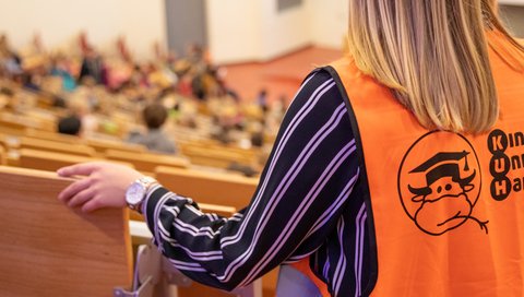 A person with an orange vest looks down from above in a lecture hall. The KinderUni logo is on the vest. 