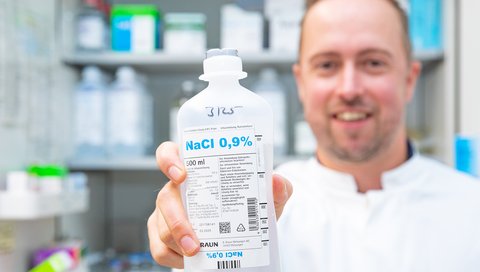 Zu sehen ist eine Flasche Natriumchlorid-Infusionslösung, die von Professor Lachmann in Richtung Kamera gezeigt wird. Professor Lachmann will die Sicherheit von Medikamenten, die durch eine Injektion parenteral verabreicht werden, revolutionieren