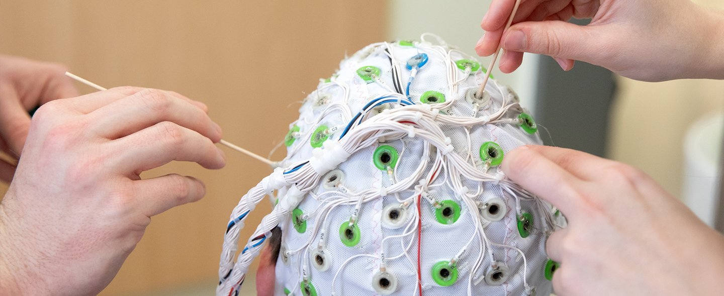 Zwei Händepaare tragen Kontaktgel auf die Elektroden einer EEG-Haube auf.
