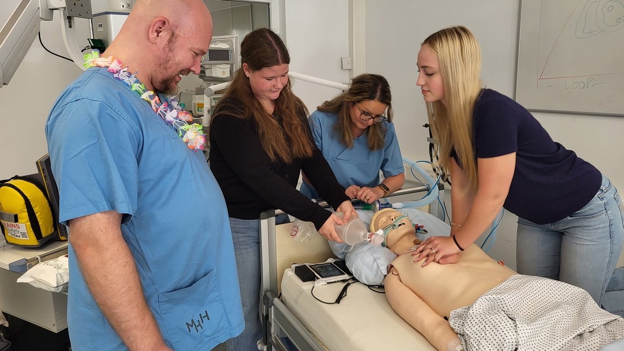 Schülerinnen üben unter Anleitung die Reanimation an einer Puppe auf einem Patientenbett. 