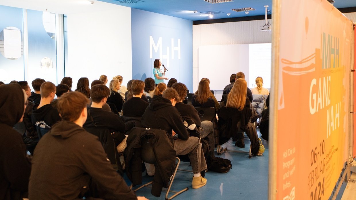 In the blue plenum at aufhof, people sit in rows of chairs and listen to a lecture. 