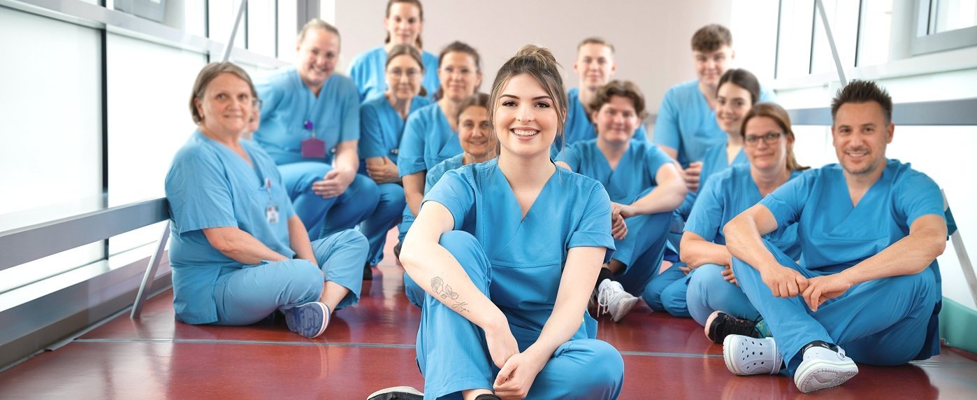 Mehrere Pflegekräfte sitzen auf dem Boden eines Flurs, ganz vorne sitzt Pflegekraft Lea und lächelt.