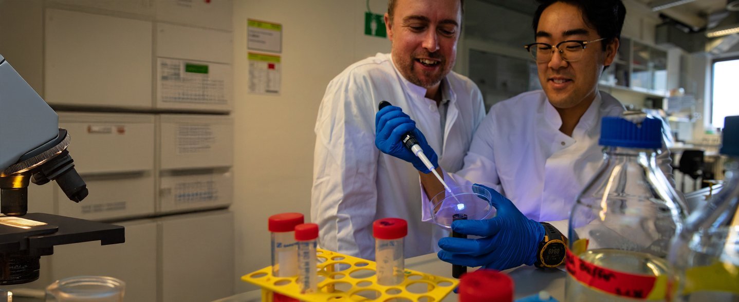 Zwei Männer in weißen Kitteln stehen und einer von ihnen hält eine Pipette in der einen und eine Schale in der anderen Hand. 