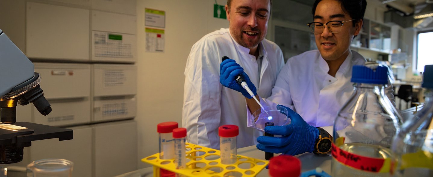Two men in white coats are standing, one of them holding a pipette in one hand and a dish in the other.