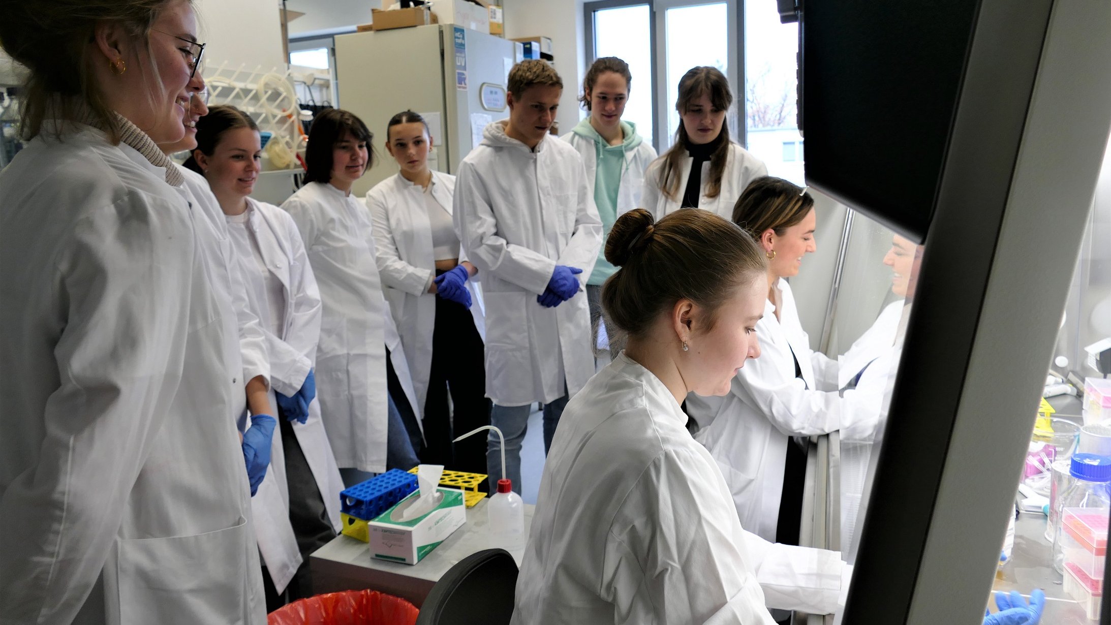 Mehrere junge Menschen stehen in weißen Kitteln in einem Labor und schauen einer anderen Person zu, die an einem Tisch ein Experiment durchführt. 