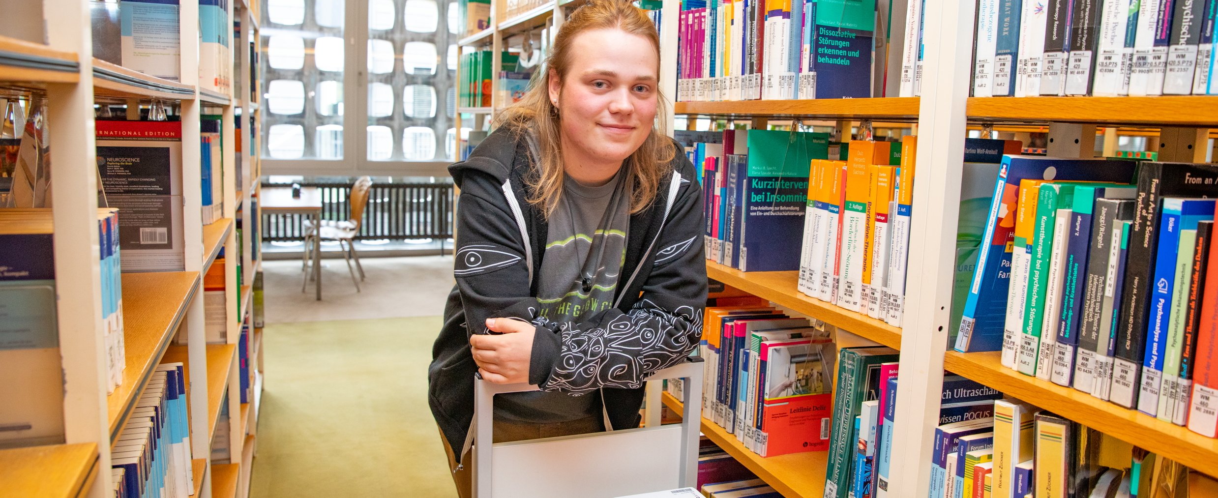 [Translate to Englisch:] Paul Vinke steht in einem Gang der MHH-Bibliothek mit Bücherregalen.