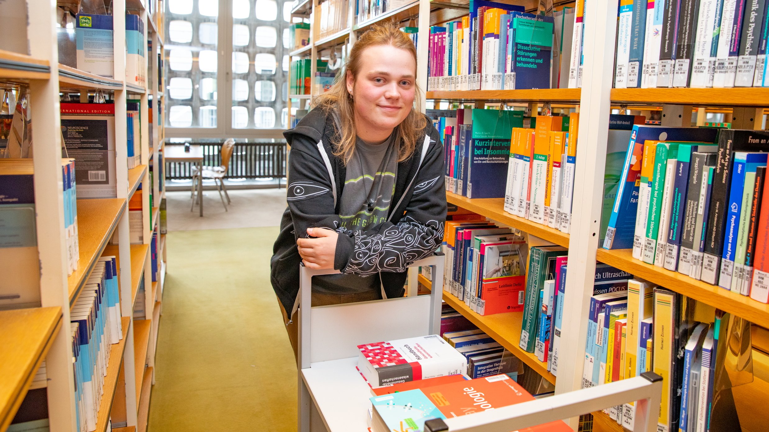 Paul Vinke steht in der Bibliothek angelehnt an einen Bücherwagen