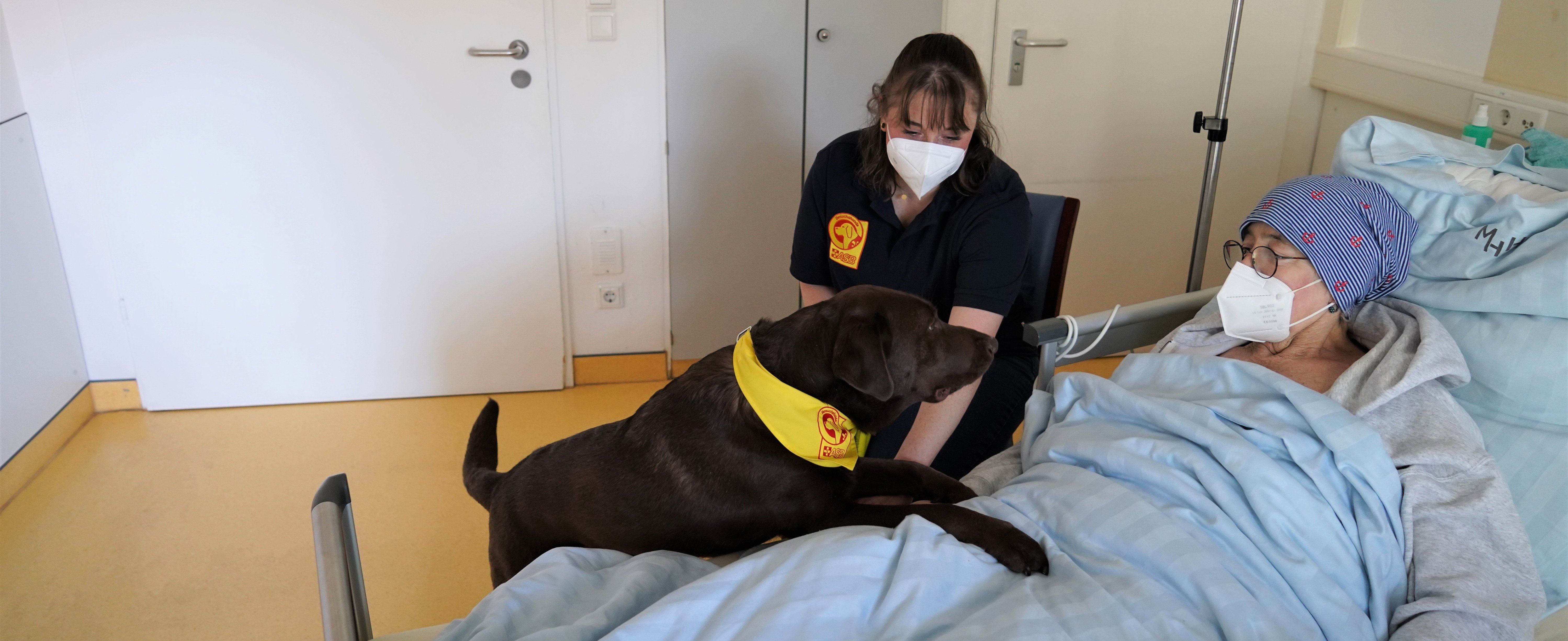 Madita Krauße sitzt mit ihrem Hund am Bett einer Palliativpatientin. Der Hund hat die Vorderpfoten auf dem Bett.